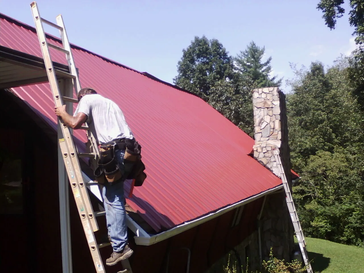 Roofer on a ladder beside a red metal roof and stone chimney — Soffit & Fascia Repair by Roofing Brothers in Mt Baldy